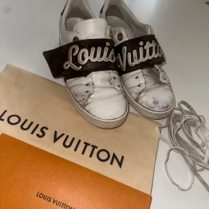Louis Vuitton vita sneakers med monogram - Snygga vita sneakers från Louis Vuitton i skinn med breda bruna monogramremmar över vristen där det står 'Louis Vuitton' i vitt. Guldiga detaljer och klassisk LV-logga baktill. Skorna har rund tå och platt sula. Medföljer originalpåse och skosnören samt kvittot. Köpta 2018 från Amager i Danmark. Äkta!!! 36,5 i storleken