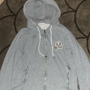 Grå Moncler hoodie med dragkedja - Snygg grå hoodie från Moncler med vit dragsko och dragkedja framtill. Tröjan har två fickor med dragkedja och broderad Moncler-logga på bröstet. Mjuk insida och huva för extra komfort. Perfekt för chill dagar eller när du vill ha en clean streetstyle.