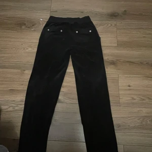 Svarta Juicy Couture mjukisbyxor - Svarta mjukisbyxor från Juicy Couture i mjuk velour med snörning i midjan och silverdetaljer. Byxorna har raka ben och broderad Juicy-logga vid benet. Perfekta för en chill och snygg look.