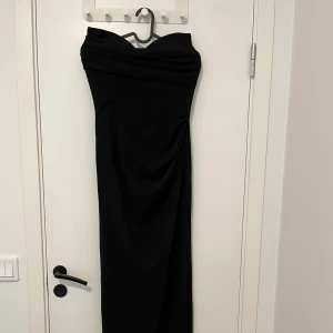 Svart axelbandslös långklänning - Elegant svart axelbandslös långklänning med draperad överdel och figurnära passform. Klänningen har en snygg, enkel design som ger en stilren och sofistikerad look. Perfekt för dig som vill sticka ut med klassisk stil.