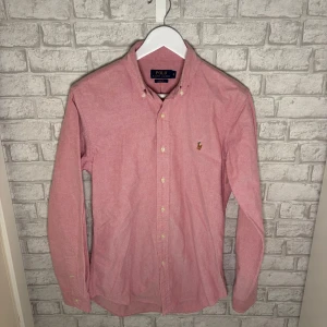 Ralph Lauren skjorta - Ralph Lauren skjorta i klassisk modell och snygg rosa färg. Stilren och lättmatchad skjorta som passar lika bra till vardag som till mer uppklädda tillfällen. I mycket bra skick, finns en liten diskret brun prick vid kragen, annars i mycket bra skick. Hör av dig vid intresse 🙌