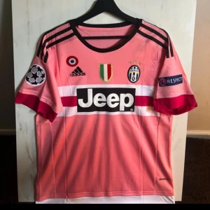 Juventus Dybala rosa fotbollströja M - Juventus fotbollströja med Dybala 21 på ryggen, tillverkad av Adidas i Climacool-material. Tröjan är rosa med svarta och vita detaljer, har korta ärmar och tryckta loggor från Champions League, Jeep och Juventus. Snygga svarta ränder på axlarna och broderade klubbmärken framtill.