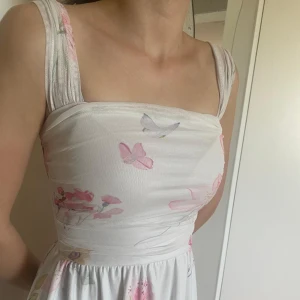 Vit långklänning med blommor - Superfin långklänning i vitt med pastellfärgade blommor och fjärilar. Klänningen har breda axelband som kan bäras både på axeln och offshoulder, samt en smickrande markerad midja och flowy kjol. Perfekt för sommarens alla tillfällen!