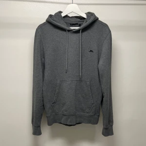 Grå hoodie från J.Lindeberg - Mörkgrå hoodie från J.Lindeberg i 100% ekologisk bomull. Klassisk modell med huva, snörning och magficka. Liten svart logga broderad på bröstet. Perfekt för chill dagar eller när du vill ha en enkel och stilren look.