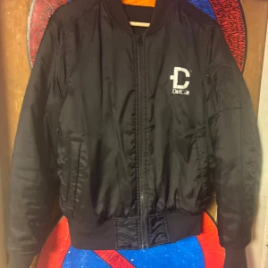 Svart bomberjacka från DirtCult - Svart bomberjacka från DirtCult med broderad logga på bröstet och orange foder. Jackan har dragkedja framtill, fickor på sidorna och en extra ficka med dragkedja på ärmen. Ribbad mudd vid ärmslut och nederkant för skön passform. Pris går att diskutera såklart