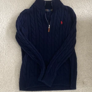 Mörkblå stickad tröja från Polo Ralph Lauren - Mörkblå kabelstickad tröja från Polo Ralph Lauren med halv dragkedja och klassisk röd broderad logga på bröstet. Tröjan har hög krage och ribbade muddar, perfekt för kyliga dagar. Materialet är mjuk ull som håller dig varm och snygg.