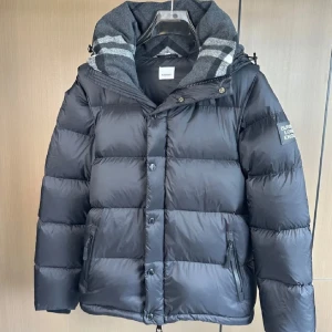 Svart dunjacka - Snygg svart dunjacka från B med klassisk rutig huva i grått och vitt. Jackan har dragkedja och knappar framtill, två fickor med dragkedja och en patch på ärmen. Perfekt för kalla dagar och riktigt stilren look. Man kan även göra den till en väst hör av er Järna 