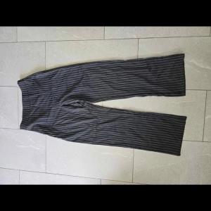 Svarta randiga kostymbyxor wide fit - Snygga svarta kostymbyxor med vita smala ränder och wide fit. Byxorna har rak passform och är perfekta för en stilren look. Materialet känns mjukt och följsamt, vilket gör dem sköna att bära hela dagen. Passar dig som gillar klassisk stil med en modern twist.