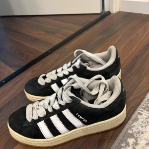 Adidas Campus svarta sneakers mocka - Säljer ett par klassiska Adidas Campus sneakers i svart mocka med vita läderdetaljer och de ikoniska tre ränderna på sidorna. Skorna har grå snörning, beige mellansula och gummisula. Perfekta för dig som gillar retrostil och streetwear.