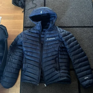 Mörkblå dunjacka Peak Performance - Säljer en mörkblå dunjacka från Peak Performance med huva och dragkedja. Jackan har quiltad design och logga på bröstet samt ärmen. Perfekt för kyliga dagar och riktigt snygg passform. Den är gjord k Sverige och är perfekt för svenska vintrar, den är helt perfekt skick och har inte som helst hål eller märken