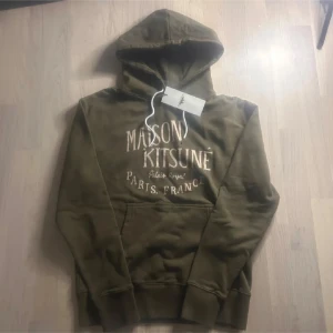 Olivgrön hoodie Maison Kitsuné  - Olivgrön hoodie från Maison Kitsuné med vit logotyptext på bröstet. Klassisk känguruficka, vita dragsnören i huvan och mjukt material. Perfekt för dig som gillar streetstyle och vill ha något snyggt och bekvämt. Nypris 4 tusen! Hör av er vid minsta fråga