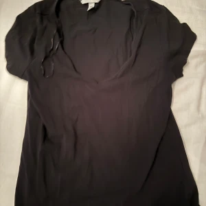 Svart kortärmad topp från H&M - En svart, enkel och stilren topp från H&M i storlek M. Toppen har korta ärmar och en djupare rundad ringning framtill. Tillverkad i mjukt och lätt material, perfekt för en clean och avslappnad look.