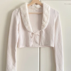 Vit croppad kofta med fuskpäls  - Superfin vit croppad kofta med ribbad struktur och fluffig fuskpäls runt kragen. Knytning framtill och långa ärmar. Perfekt för dig som vill ha en söt och trendig look med lite extra detaljer 🤍