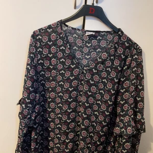 Blommig svart klänning från Ddlns - Svart klänning med v-ringning och blommigt mönster i rött, vitt och grönt. Lätt och luftig modell med trekvartsärm och volangdetaljer. Perfekt för dig som gillar en romantisk och bohemisk stil.