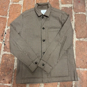 Bläck overshirt - Snygg rutig overshirt från Bläck i bruna och beige toner. Jackan har klassisk krage, knäppning framtill och en bröstficka. Perfekt lager-på-lager-plagg med stilren skandinavisk känsla.