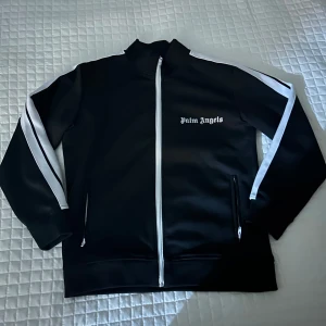 Svart track jacket från Palm Angels - Palm Angels svart track jacket med vita ränder längs ärmarna och vit dragkedja. Jackan har ståkrage, två fickor med dragkedja och logotypen tryckt på bröstet. Materialet är polyester och modellen är loose fit med långärmade ärmar.
