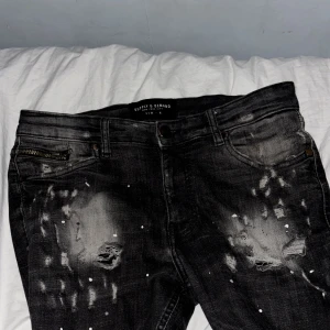 Svarta slitna jeans med stänk och hål - Säljer ett par svarta jeans med grå tvätt, slitningar och hål på flera ställen samt vita färgstänk. Jeansen har klassisk femficksdesign och raka ben. Perfekt för dig som gillar en edgy och trendig look.