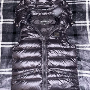 Svart dunväst från Bvaldi - Snygg svart dunväst från Bvaldi med glansig finish och hög krage. Västen har två dragkedjefickor framtill, avtagbar huva med tryckknappar och ett stilrent Bvaldi-märke på bröstet. Perfekt för lager-på-lager under kyliga dagar.
