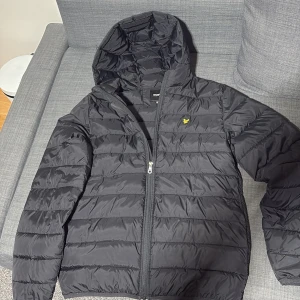 Svart pufferjacka från Lyle & Scott - Svart pufferjacka från Lyle & Scott med huva och dragkedja. Jackan har quiltad design och en gul logga på bröstet. Perfekt för kyliga dagar och har en clean, sportig look. Passar dig som gillar stilrena och funktionella plagg.
