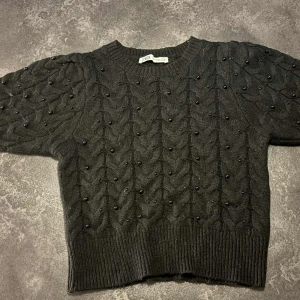 Svart stickad tröja med pärlor Zara - Supersnygg svart stickad tröja från Zara med korta ärmar och dekorativa svarta pärlor över hela tröjan. Tröjan har ett flätstickat mönster och ribbade muddar vid hals, ärmslut och nederkant. 