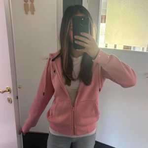 Rosa hoodie med dragkedja och huva - Säljer en rosa hoodie med dragkedja framtill och huva med snörning. Tröjan har två fickor på magen och ribbade muddar vid ärmslut och nederkant. Perfekt för en chill och avslappnad stil.