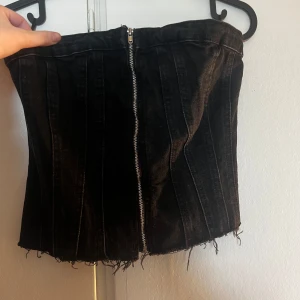 Svart jeanskorsett från Zara - Säljer en svart jeanskorsett från Zara med dragkedja framtill och råa, fransiga kanter nertill. Toppen är axelbandslös och har markerade sömmar för en snygg siluett. Baksidan är smockad för extra komfort och passform. Perfekt för en edgy look!
