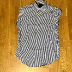 Randig skjorta från Lauren Ralph Lauren - Snygg skjorta från Lauren Ralph Lauren med blå och vita ränder. Klassisk krage, långärmad och knäppning framtill. Tillverkad i 100% bomull för en skön känsla. Enbart testad en gång, köpt sprillans ny. Ångrade köpet men slarvat bort kvittot. 
