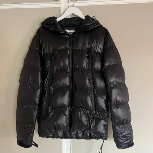 Svart pufferjacka från Gentlemen's Corner - Svart pufferjacka med huva från Gentlemen's Corner. Jackan har en glansig finish, två vertikala dragkedjefickor framtill och justerbara muddar. Perfekt för kalla dagar med sin vadderade design och snygga, sportiga look.