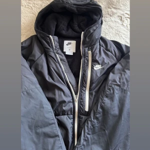Svart Nike pufferjacka med huva - Säljer en svart pufferjacka från Nike med huva och vit dragkedja. Varm, bekväm och vindtät med fleecefoder på insidan. Perfekt för kalla dagar och sportig vardagsstil.