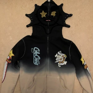Glo Gang hoodie (Chief Keef Merch) - Unik Glo Gang hoodie med grymt cool stjärnmask på huvan, mörkblå/svart och beige färgton med snygg gradient. Tryck med dödskalle, stjärnor och texten 'Death and Glory' samt 'the sun will rise!' på ryggen. Dragkedja framtill och två fickor. Perfekt statement-plagg för dig som vill sticka ut.
