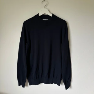 Mörkblå stickad tröja från Selected Homme - En stilren mörkblå stickad tröja från Selected Homme med ribbad krage och muddar. Tröjan har en klassisk passform och är tillverkad i en mix av bomull och återvunnen polyester. Perfekt för lager-på-lager under hösten och vintern. 2M2 