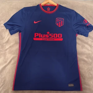 Atlético Madrid blå Nike fotbollströja - Snygg blå Atlético Madrid fotbollströja från Nike med röda detaljer och klubbmärke på bröstet. Tröjan har korta ärmar, rund hals med röda ränder och tryckta sponsorer. Tillverkad i lätt och ventilerande material, perfekt för match eller träning. pris kan diskuteras