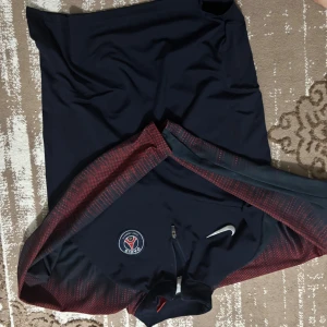 PSG långärmad träningströja Nike - Mörkblå långärmad fotbollströja från Nike med PSG-logga på bröstet och halv dragkedja. Ärmarna har röda detaljer i ett diskret mönster. Tillverkad i ett lätt och sportigt material som andas, perfekt för träning eller match.