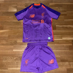 Liverpool Salah fotbollsset lila - Liverpool FC fotbollströja och shorts i lila med orange detaljer, tryckta loggor och sponsor. Tröjan har korta ärmar, M. Salah och nummer 11 på ryggen. Tillverkad i lätt och ventilerande funktionsmaterial, perfekt för träning eller match.