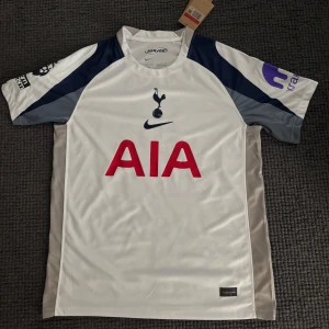 Tottenham Hotspur Kudus 20 Nike tröja - Säljer en Tottenham Hotspur fotbollströja från Nike med Kudus och nummer 20 på ryggen. Tröjan är vit med marinblå och grå detaljer, röd AIA-logga på bröstet och Premier League-märke på ärmen. Tillverkad i Dri-FIT-material för bästa komfort.