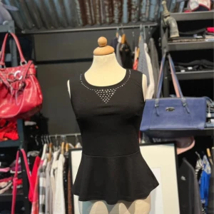 Svart peplumtop med nitar  - Svart ärmlös peplumtop med rund halsringning och dekorativa svarta nitar i ett triangulärt mönster framtill. Toppen har figurnära passform upptill och utsvängd nederdel. Perfekt för dig som gillar edgy detaljer och stilren look.