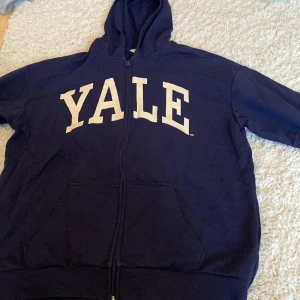 Mörkblå Yale hoodie från H&M - Säljer en mörkblå hoodie från H&M med stor vit YALE-text på bröstet. Tröjan har dragkedja framtill, huva och två fickor. Perfekt för dig som gillar college-stil och vill ha något bekvämt och snyggt till vardags.