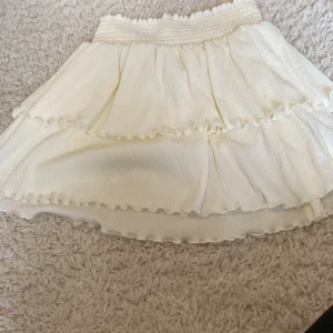 Vit volangkjol från Kappahl - Supersöt vit kjol från Kappahl med bred resår i midjan och dubbla volanger. Kjolen har en lätt rynkad struktur och vågiga kanter som ger en extra fin touch. Perfekt för dig som gillar en luftig och feminin stil.