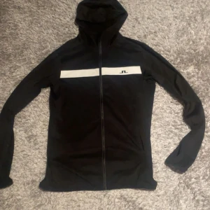 Svart kofta från J.Lindeberg - Svart kofta från J.Lindeberg med vit horisontell rand och logga på bröstet. Tröjan har dragkedja framtill, huva och långa ärmar. Perfekt för en sportig och stilren look.