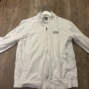 EA7 Armani tröja kofta - Snygg vit ziptröja från EA7 Emporio Armani med hög krage och diskret logga på bröstet. Tröjan har hel dragkedja, två fickor och är tillverkad i mjuk bomull. Perfekt för en clean och sportig look. använd en gång, L sitter som medium