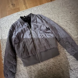 Grå quiltad bomberjacka - Säljer en grå bomberjacka med quiltade ärmar och ribbade muddar. Jackan har dragkedja framtill och två snedställda fickor. Perfekt för dig som gillar en clean och stilren look.