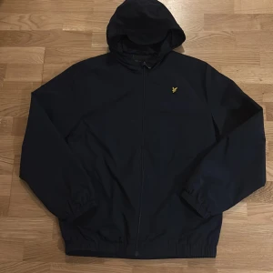 Svart vindjacka från Lyle & Scott - Svart vindjacka från Lyle & Scott med huva och dragkedja framtill. Jackan har det klassiska gula logobroderiet på bröstet och elastiska muddar vid ärmslut och midja. Tillverkad i polyester och har en stilren, enkel design som passar perfekt till streetwear.