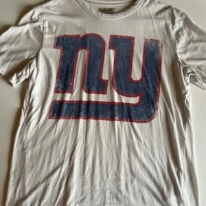 Vit Giants t-shirt från Jack & Jones - Snygg vit t-shirt från Jack & Jones VintageClth med stort blått och rött NY-tryck fram och GIANTS-tryck bak. Klassisk rund hals och korta ärmar. Perfekt för dig som gillar sportiga och retroinspirerade plagg.