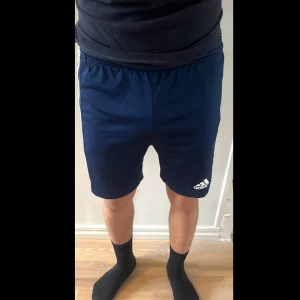 Blå Adidas shorts med vita ränder - Snygga blå träningsshorts från Adidas med klassiska vita ränder längs sidorna och Adidas-logga på ena benet. Shortsen har elastisk midja och är gjorda i ett lätt och svalt polyestermaterial, perfekta för sport eller chill. Passar dig som gillar sportig stil. Storlek: 13-14 år (164)
