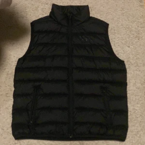 Svart dunväst från Kappahl - Svart dunväst från Kappahl med hög krage och dragkedja framtill. Västen har quiltad design och två fickor med dragkedja. Perfekt för lager-på-lager och enkel att matcha med olika outfits. Jag har nästan aldrig använt den, köptes på killavdelningen men tycker att tjejer också kan ha den. 