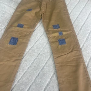 Beiga chinos med blå prickiga lappar - Unika beiga chinos från Guess med blå prickiga lappar och synliga gula stygn på benen. Byxorna har raka ben, bälteshällor och blåprickigt tyg även på insidan av bensluten. Perfekt för dig som vill sticka ut med en kreativ och personlig stil.