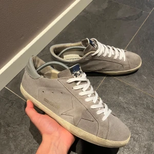 Gråa Golden Goose - Snygga gråa Golden Goose Superstar sneakers i mocka med vit sula och vita skosnören. Skorna har GGDB-logga och palmmotiv på plösen samt den ikoniska stjärnan på sidan. Perfekta för dig som gillar en clean och stilren look.