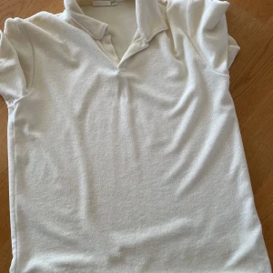 Vit frotté t-shirt från Kappahl - Vit t-shirt från Kappahl i mjuk frotté med korta ärmar och klassisk krage. Enkel och clean look, perfekt för dig som gillar retrovibbar och sköna material. Snyggt avslappnad passform och stilren design.