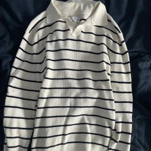 Randig pikétröja i bomull - Vit pikétröja med svarta horisontella ränder från Volt. Tröjan har klassisk krage, lång ärm och är tillverkad i ett strukturerat bomullstyg. Perfekt för en stilren och avslappnad look.