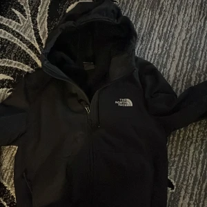 Svart vindjacka från The North Face - Säljer en svart vindjacka från The North Face med huva och dragkedja framtill. Jackan har en enkel och stilren design med logga på bröstet och en ficka med dragkedja. Perfekt för blåsiga dagar och passar till det mesta.
